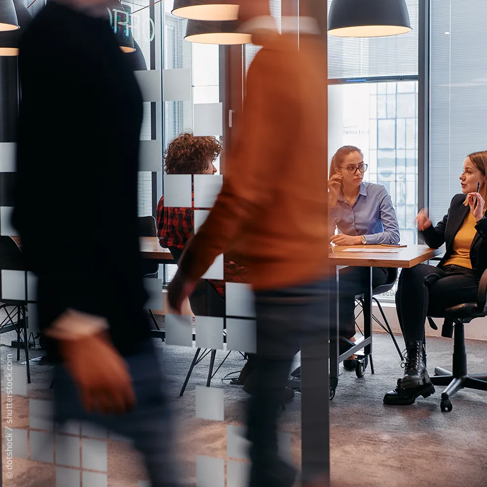 Besprechung im Hintergrund währen im Vordergrund Mitarbeiter durchs Office laufen